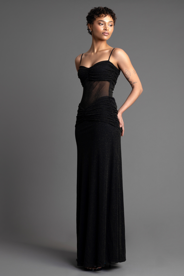 model wearing a black glittery maxi dress with spaghetti straps, ruched bust, sheer midsection, and a floor-length skirt