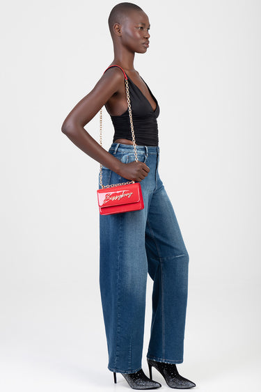 model in a black ruched top and blue wide leg jeans carrying a red crossbody bag with a gold chain strap
