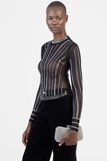 woman in a sheer black long sleeve top with white dotted lines and a high neckline, paired with black pants and a beaded