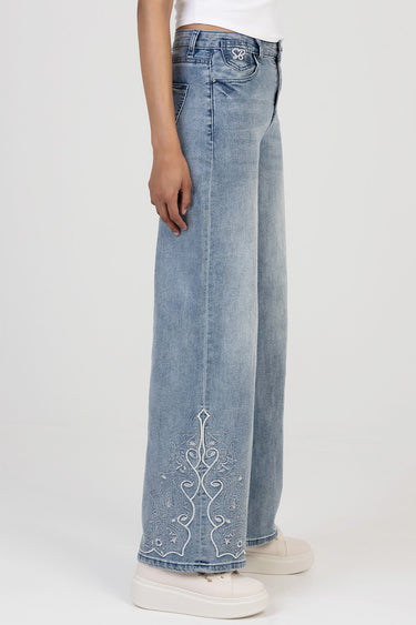 side view of a woman wearing light blue wide leg jeans with white embroidered detailing at the lower leg and white sneakers