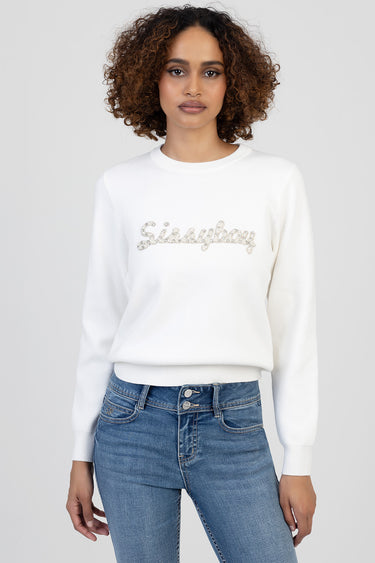 woman wearing white knit sweater with silver sequin lettering across the chest and blue jeans
