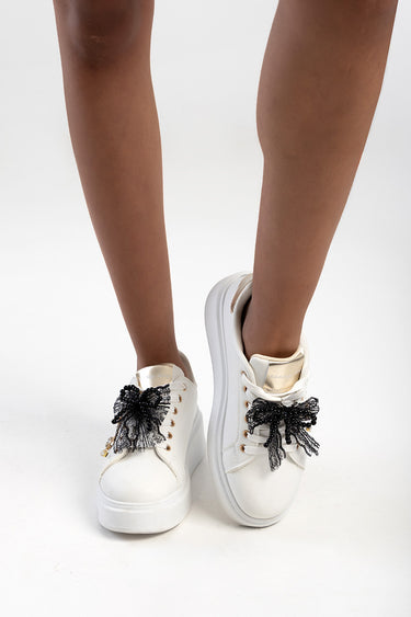 Blue Jean Traders white sneakers with black lace bow decorations on the laces and a metallic gold tongue.