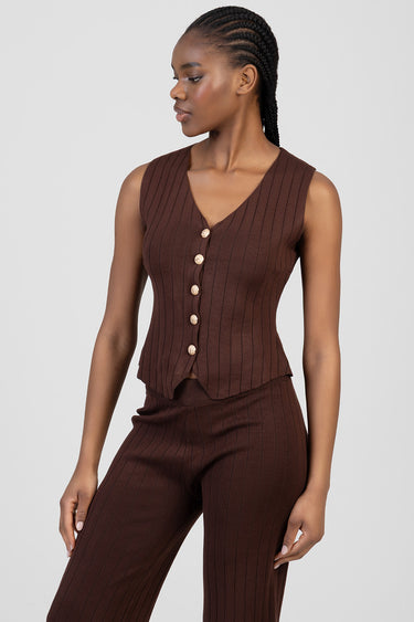 woman wearing a brown ribbed sleeveless waistcoat with a v neck and gold buttons, paired with matching brown ribbed