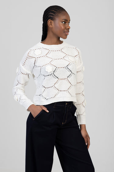 woman wearing a white open knit sweater with circular lace pattern and flower appliques, paired with dark wide leg trousers