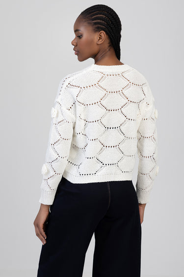 woman wearing white knit sweater with open scalloped pattern, back view, pom pom accents on sleeves, dark trousers