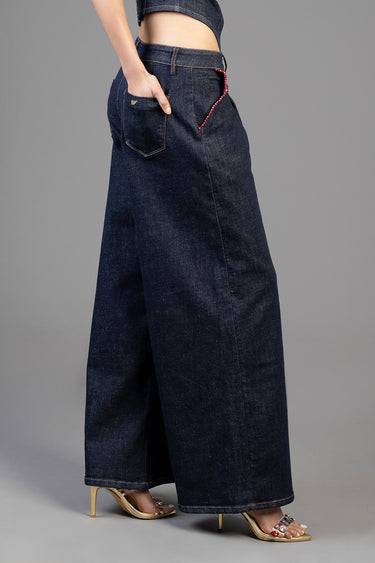 woman in dark blue wide leg jeans with large back pockets and a red beaded chain detail, standing in gold heeled sandals