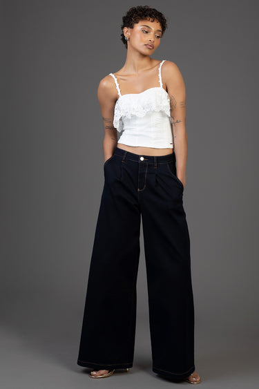 woman in white lace strap top with a ruffled overlay and wide black denim trousers, hands in pockets, grey studio backdrop