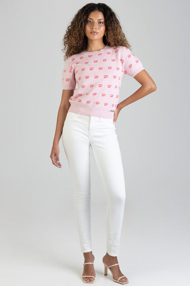 woman in a light pink short sleeve knit top with red bow motifs and white skinny jeans, beige heeled sandals, standing pose