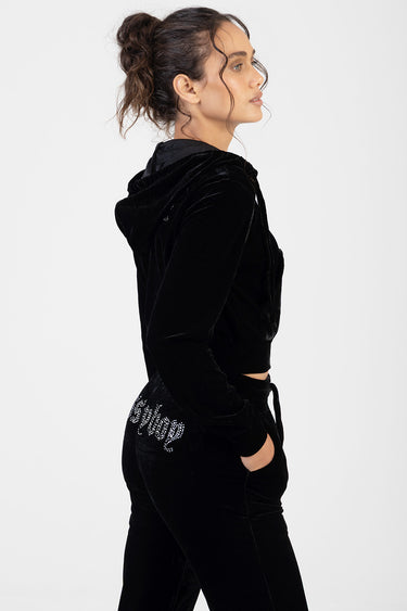 woman wearing a black velour cropped hoodie and matching joggers with a rhinestone logo on the hip