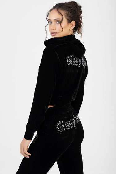 woman wearing black velour tracksuit with hoodie, rhinestone script on back and hip, standing turned to the side