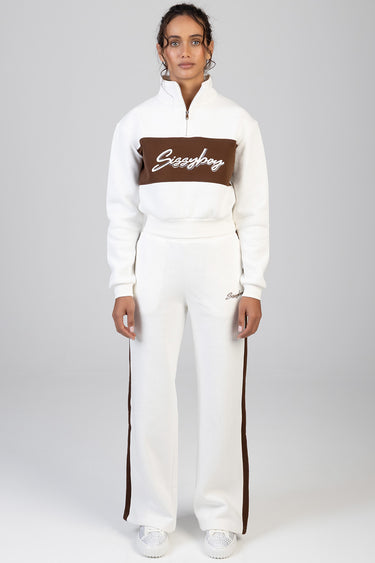 woman models a white track set with a brown chest band and brown side stripes, logo script on chest and pant leg, white