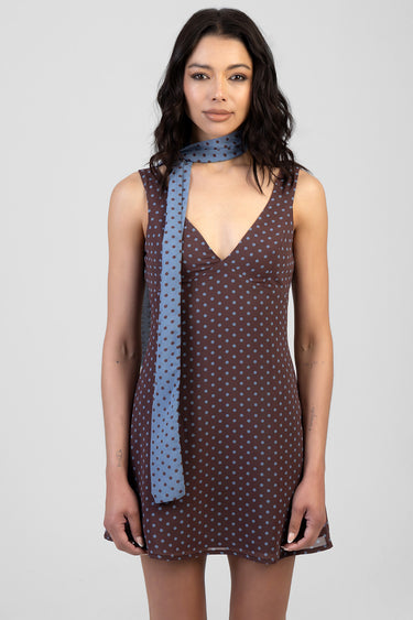woman wearing a brown sleeveless polka dot mini dress with a light blue scarf around the neck