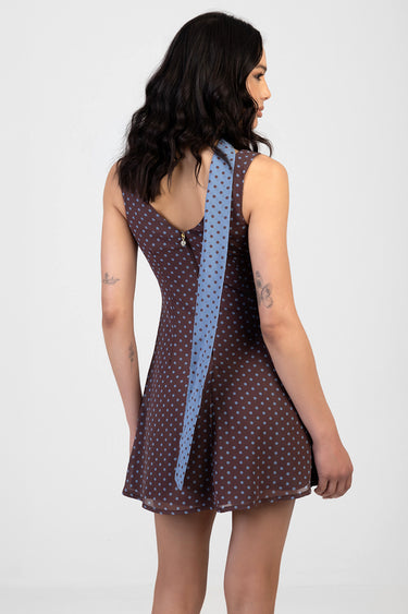 woman wearing a brown sleeveless polka dot mini dress with a light blue scarf draped down the back