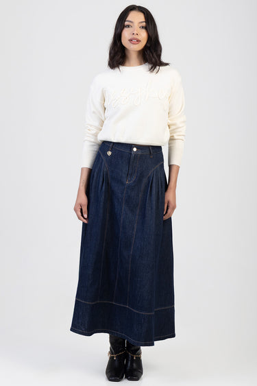 woman in a cream knit sweater and a long dark denim skirt with pockets and visible stitching, black ankle boots with a gold