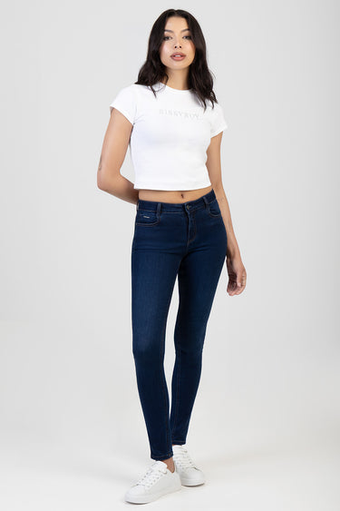 woman wearing dark blue skinny jeans with a white cropped t shirt and white sneakers standing against a light background
