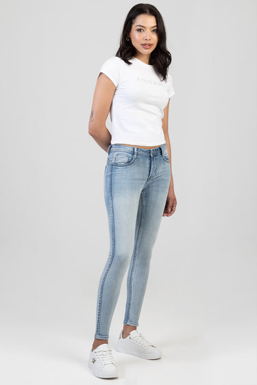 woman wearing light blue skinny jeans with a white t shirt and white sneakers, standing against a plain backdrop