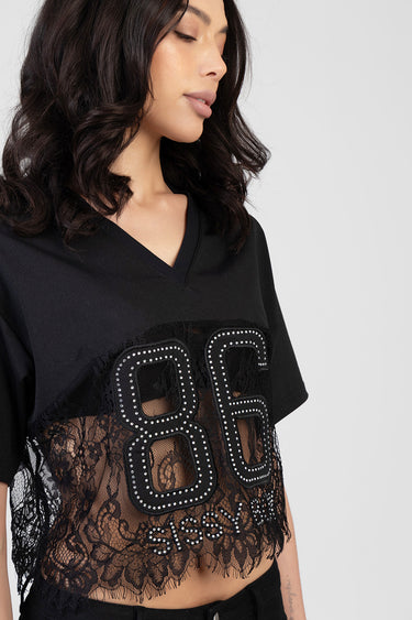 woman wearing a black boxy v neck top with sheer lace panel and silver studded eighty six design across front
