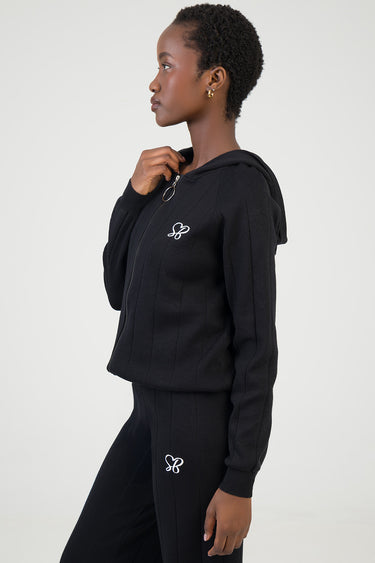 side view of a woman wearing a black knit zip hoodie with hood and matching black knit pants with small white logos