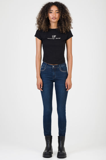 woman wearing dark blue skinny jeans, black logo t shirt, and black ankle boots, standing against a light background