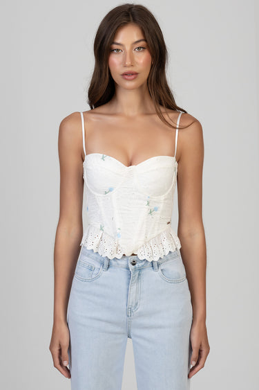 woman wearing a white corset top with floral embroidery and a scalloped lace hem, thin straps, paired with light wash jeans