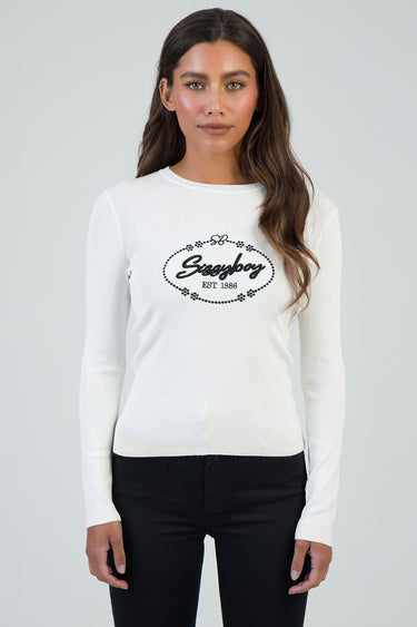 woman wearing white knit long sleeve top with a black oval logo and floral border across the chest