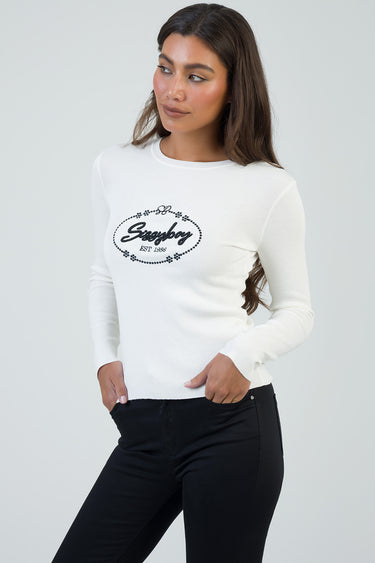 white knit long sleeve top with a black circular logo on the chest, paired with black jeans