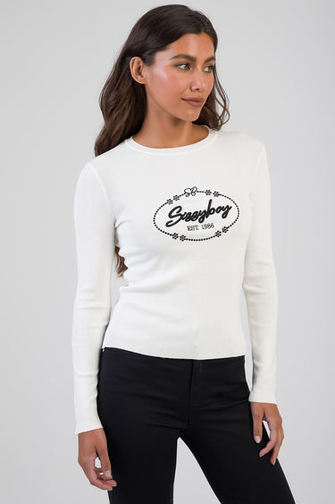 white knit top with black oval logo and floral border, long sleeves, worn by a model in black jeans