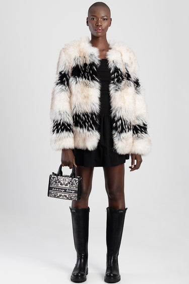 woman in a black mini dress and striped cream and black faux fur coat, carrying a patterned mini handbag with tall black