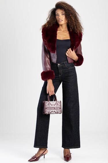 a woman wearing a burgundy fur trim jacket over a black top and dark jeans, holding a small patterned handbag.