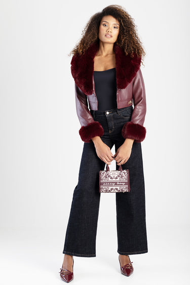 woman in a burgundy faux leather jacket with fur collar and cuffs, wide dark jeans, holding a small patterned maroon handbag