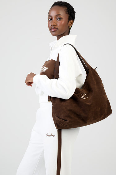 woman in white athleisure carries a brown suede tote bag by Blue Jean Traders with a logo on the side