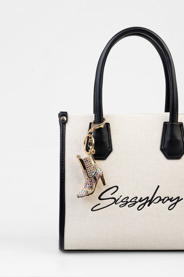 beige canvas handbag with black leather trim and handles, gold hardware, and a jeweled boot charm hanging from a ring