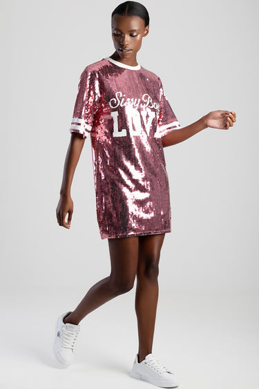 woman wearing a pink sequin t shirt dress with white trim and large white lettering, white sneakers