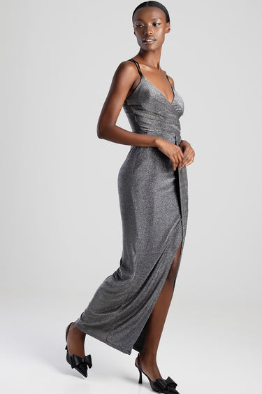 model wearing a silver metallic maxi dress with thin straps, wrap at the waist, and a front slit, paired with black heeled