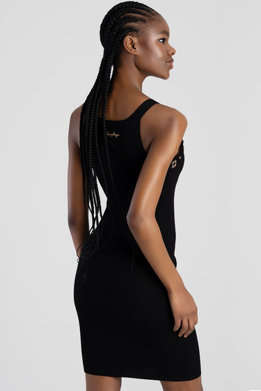 woman wearing a black knit sleeveless dress, viewed from behind, with long braids and a small gold logo on the back.