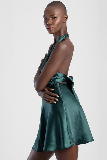 woman in emerald green satin halter dress with open back and bow at the waist, standing in profile