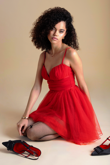 woman wearing a red spaghetti strap tulle mini dress sits on the floor with fishnet tights and black embellished heels nearby