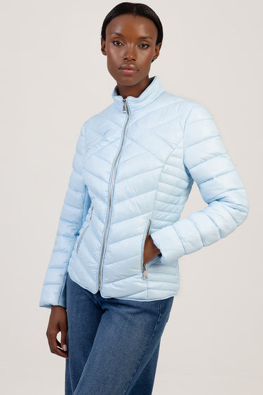 woman wearing a light blue puffer jacket with a high collar and silver zip, standing against a beige backdrop