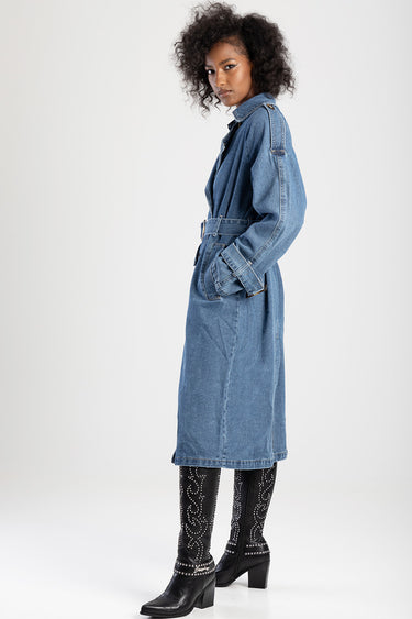 woman wearing a blue denim trench coat with a belt, standing in profile, paired with black studded knee boots