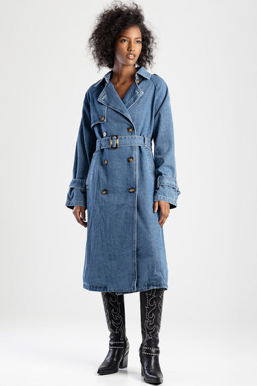 woman modeling a blue denim trench coat with a belt, double breasted buttons and cuff buckles, paired with studded black