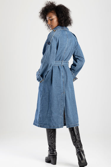 woman wearing a long blue denim trench coat with belt, back view, black studded knee boots