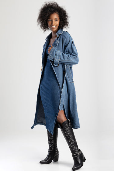 woman wearing a long blue denim trench coat belted at the waist, with black studded knee high boots
