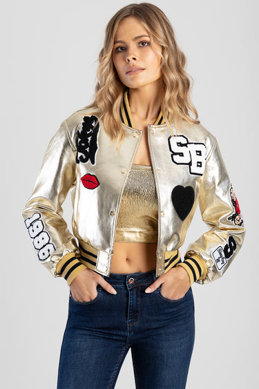 woman wearing metallic gold bomber jacket with patches and striped cuffs, paired with a gold top and dark jeans