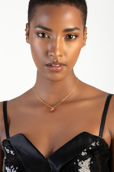 woman with short hair wearing a gold chain necklace with a red cherry pendant and small hoop earrings, black sequined top
