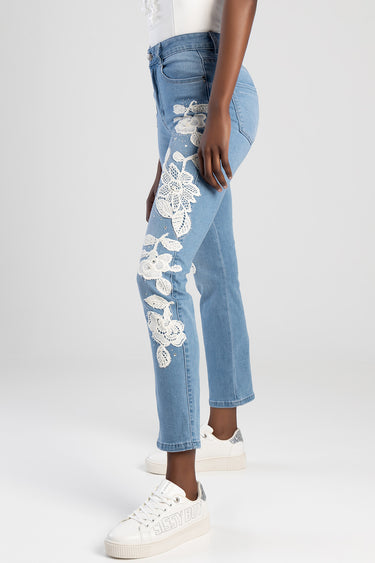 light blue denim jeans with large white floral lace appliqué along the outer leg, cropped at the ankle, shown with white
