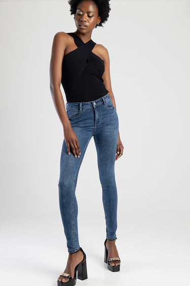 woman wearing blue skinny jeans and a black crisscross sleeveless top, standing against a light backdrop