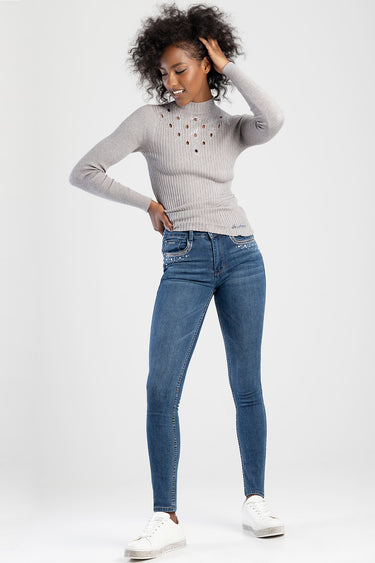woman posing in a light gray knit sweater with cutouts and slim blue skinny jeans with rhinestone accents, white sneakers