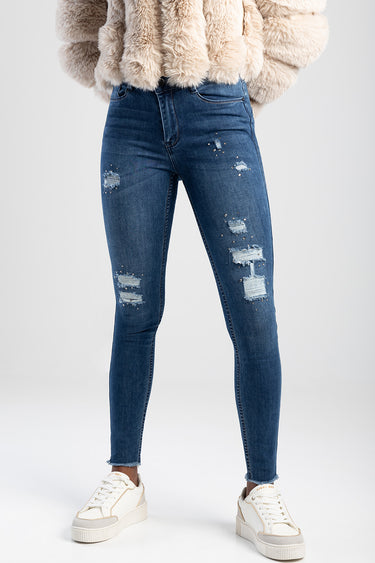 model wearing dark blue distressed jeans with ripped patches and frayed hems, paired with white sneakers