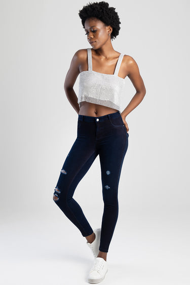 model in dark indigo ripped skinny jeans and a silver knit cropped top with wide straps, white sneakers, standing against a