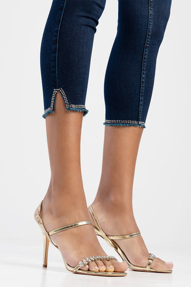 woman wearing dark blue capri jeans with rhinestone trim and frayed edges, paired with gold strappy heeled sandals.